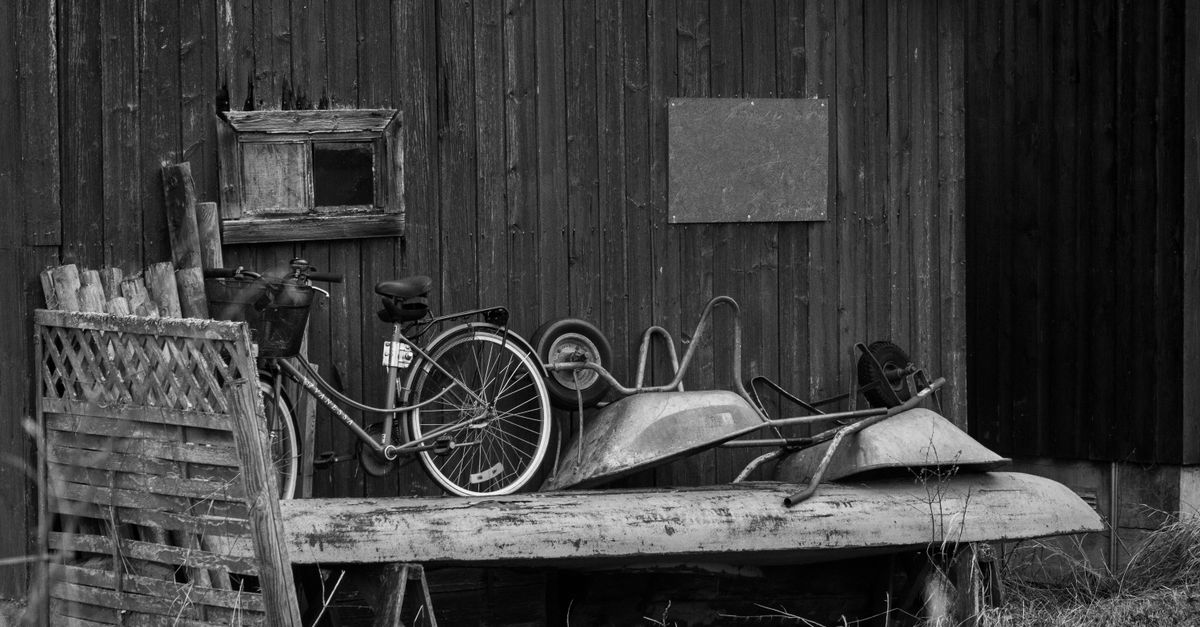 Zwart-witfoto van een oude fiets en kruiwagens met rommel voor een houten schuur