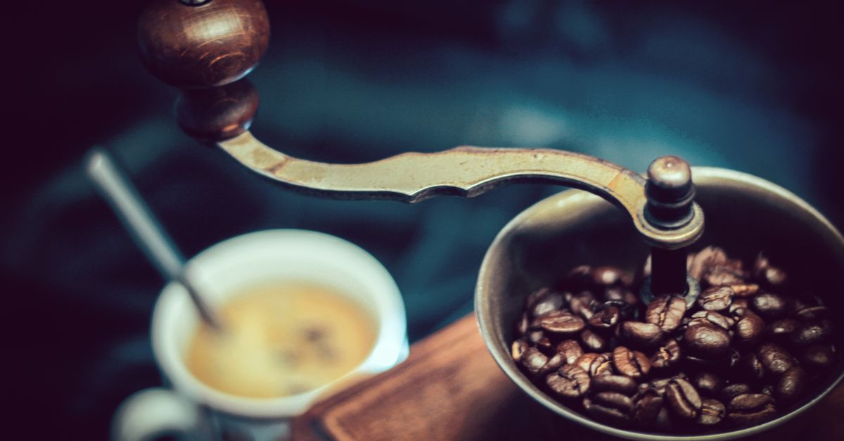 Een handmatige koffiemolen gevuld met koffiebonen naast een kopje verse koffie op tafel