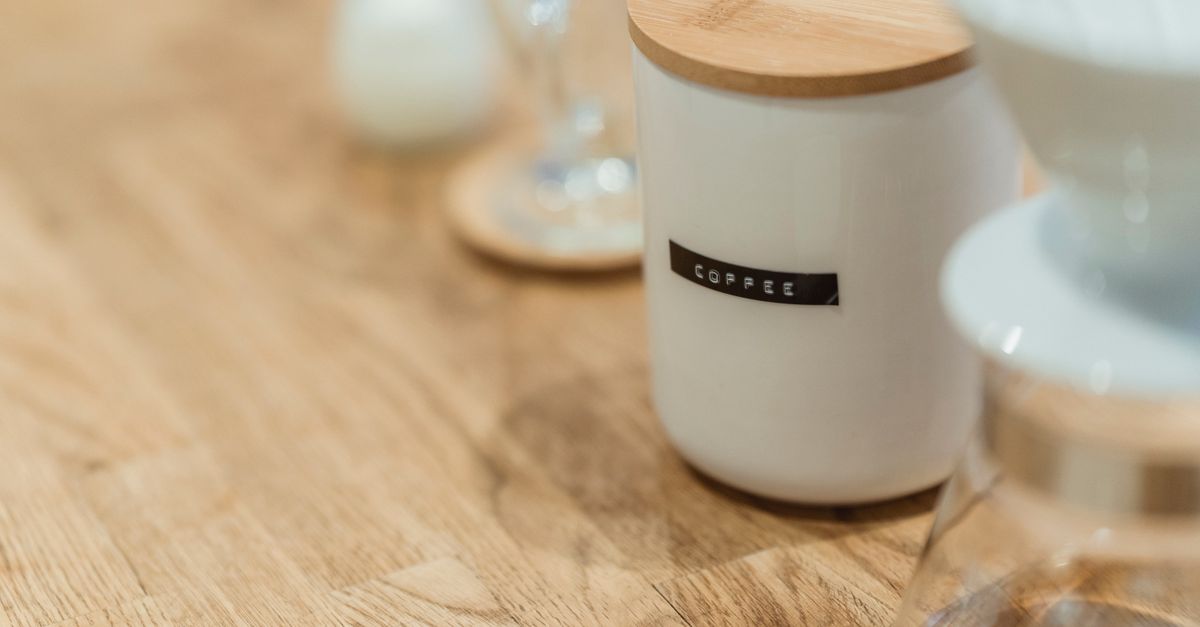 Een witte keramische koffiebus met houten deksel en het label 'coffee' op een houten aanrecht, met daarnaast een koffiefilterhouder en glazen.