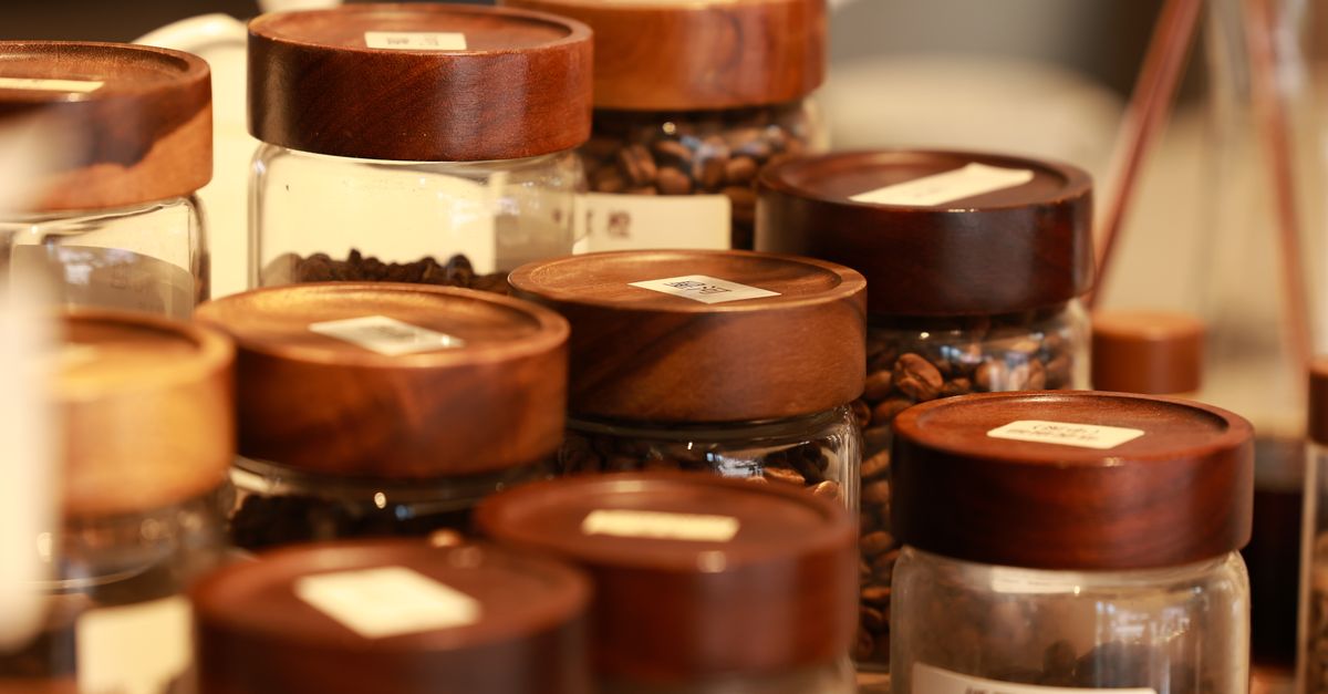 Rijen glazen potjes met houten deksels gevuld met koffiebonen, dicht bij elkaar op een plank.