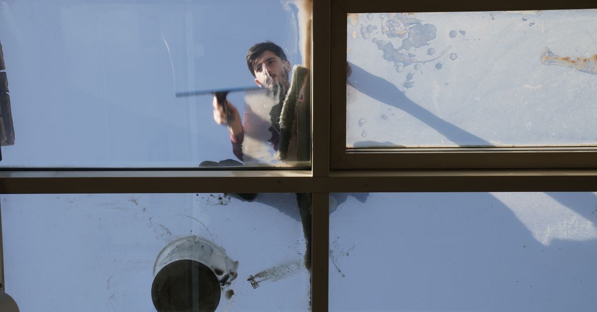 Een man wast een raam met een wisser vanaf de binnenkant, gezien door het glas met zonlicht en vlekken op het venster.