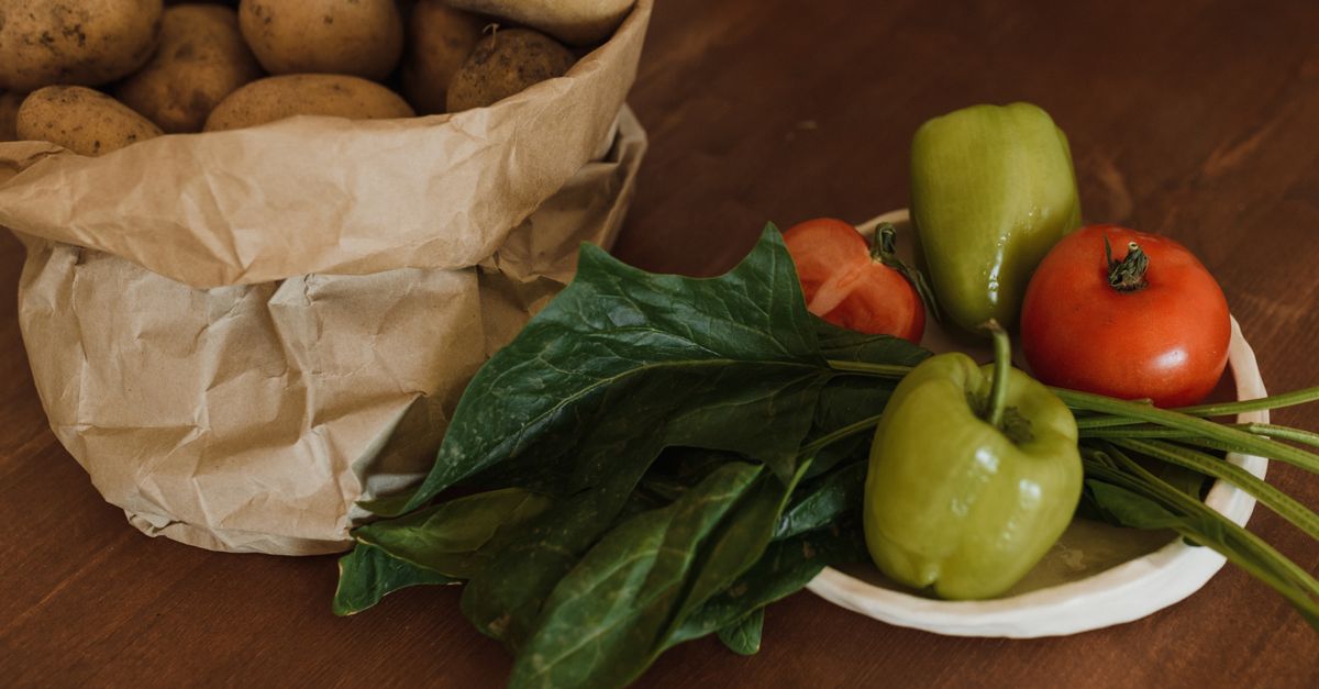 Rauwe aardappelen in een papieren zak op een houten ondergrond, naast een schaal met paprika's en tomaten.