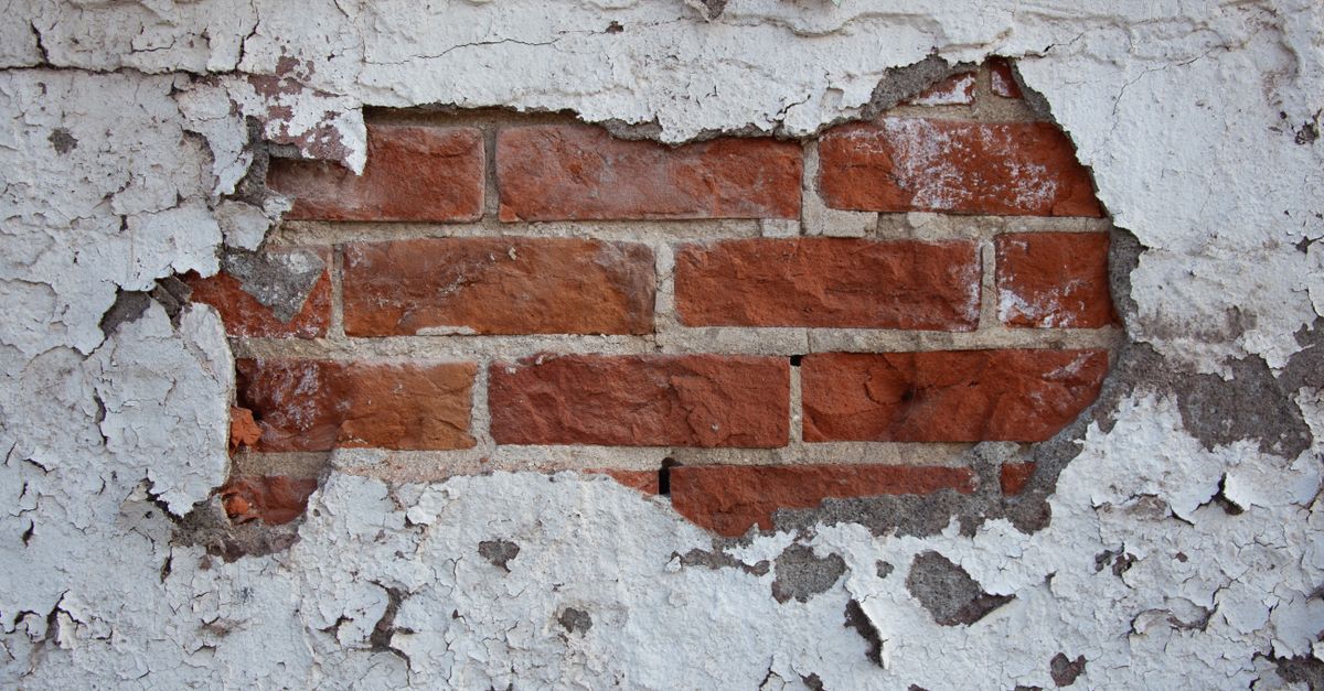 Afbladderende witte verfstructuur op een muur waar de rode bakstenen eronder zichtbaar worden door vochtschade.