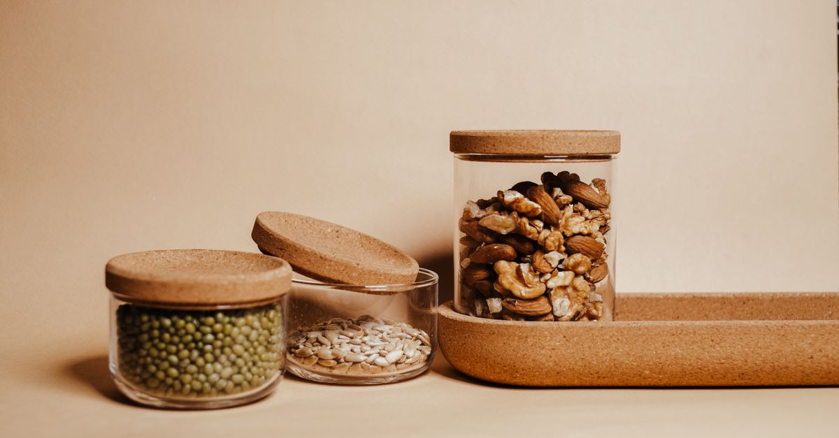 Glazen potten met kurken deksel gevuld met noten, groene mungbonen en zonnebloempitten op een beige ondergrond.
