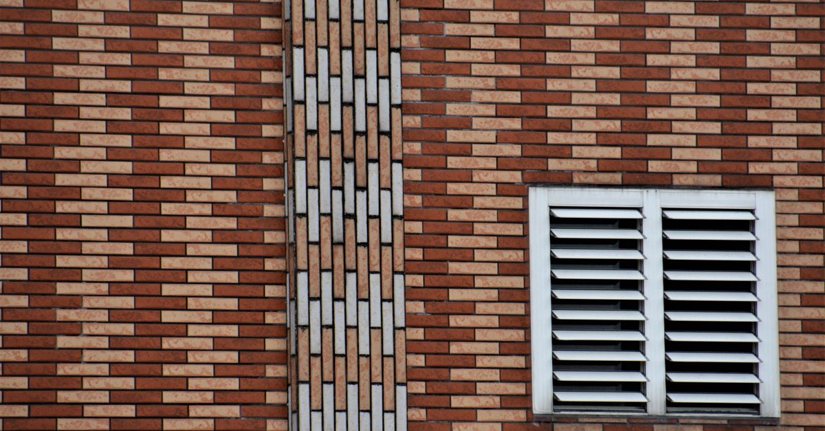 Witte ventilatierooster geplaatst in een bakstenen gevel naast een raam.