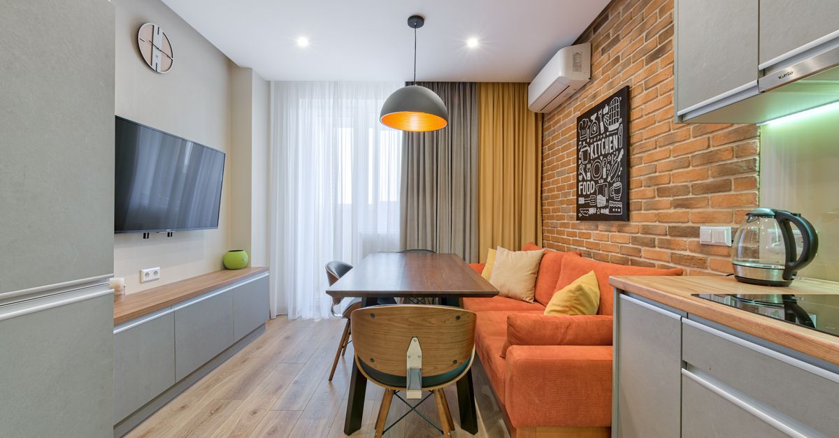 Compacte open woonkamer met keuken, oranje bank, eettafel met stoelen en een bakstenen accentwand in een klein appartement