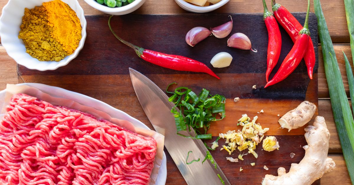 Bord met rauw gehakt naast een snijplank met knoflook, chilipepers, gember en kruiden, klaar om te bereiden.