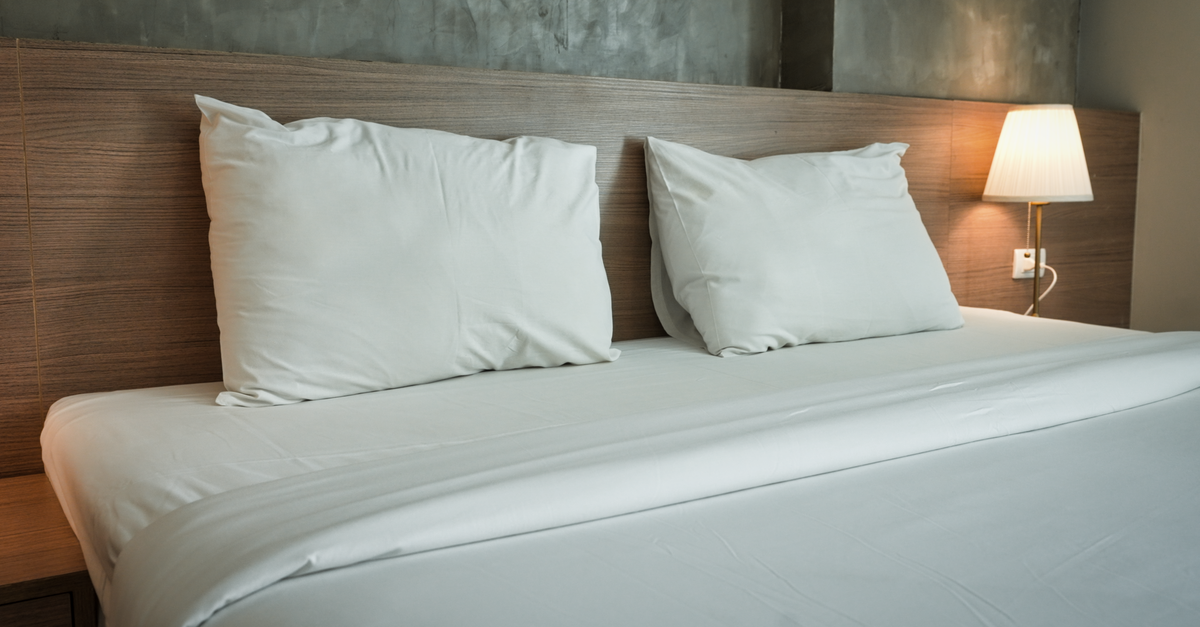 Een opgemaakt bed met wit beddengoed en twee witte kussens tegen een houten hoofdbord, met een lampje op het nachtkastje.