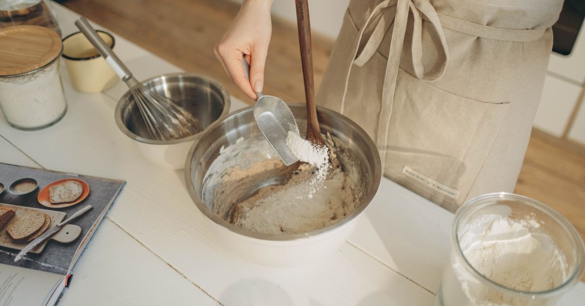Een hand giet met een lepel bloem uit een pot in een metalen mengkom, terwijl er ook een garde en een weegschaalpot met bloem op het aanrecht staan.