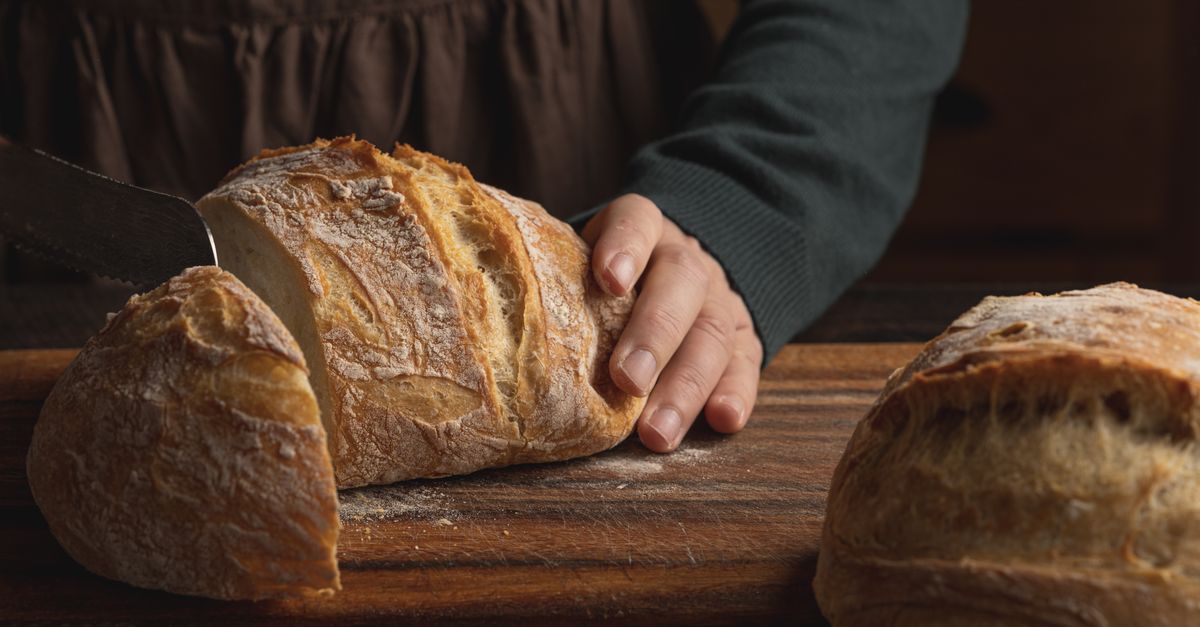Close-up van een hand die een knapperig brood snijdt met een mes op een houten snijplank.