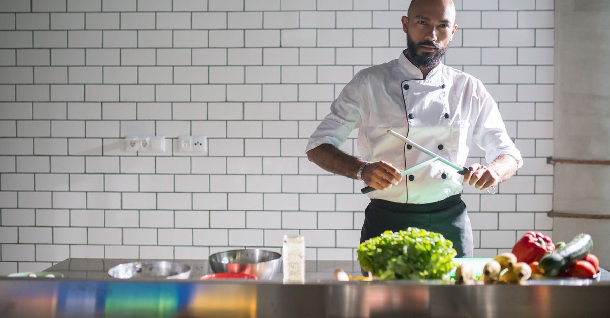 Een chef-kok in koksbuis wet een mes met een aanzetstaal boven een keukenwerkblad met groenten en kookgerei.