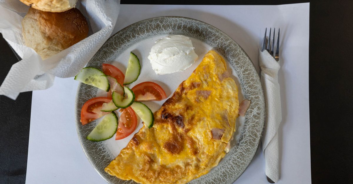 Een gevouwen omelet met ham op een grijs bord, geserveerd met verse tomaat, komkommer en een schepje roomkaas.
