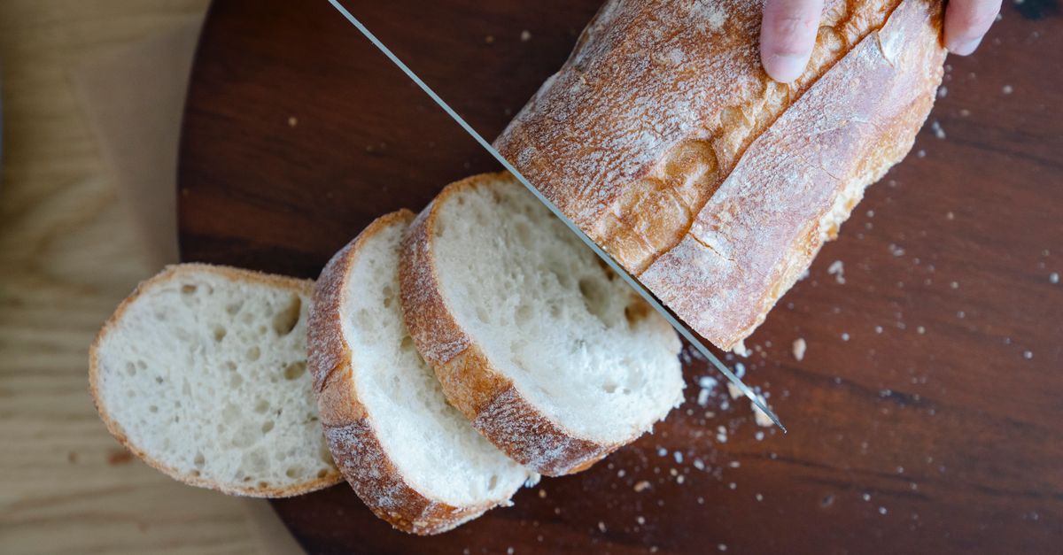 Een hand snijdt met een mes vers brood in plakken op een ronde houten snijplank.