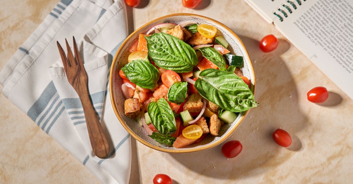 Een kom panzanella met broodblokjes, tomaten, komkommer en grote basilicumblaadjes, geserveerd op een keukendoek met vork en lepel.