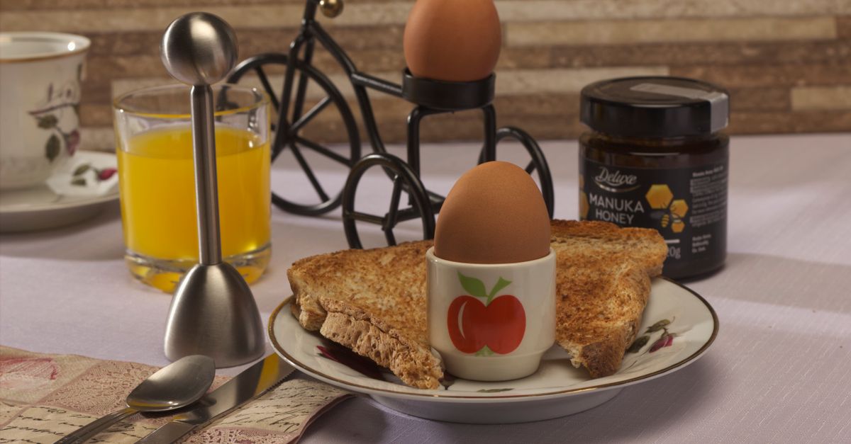 Een zachtgekookt eitje in een eierdopje met appelmotief, geflankeerd door geroosterd brood, sinaasappelsap en een pot honing op tafel.