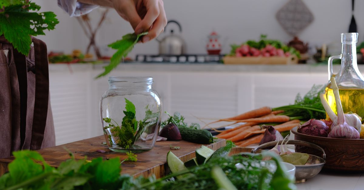 Een hand stopt verse groene kruiden in een glazen pot, omringd door groenten zoals wortels, knoflook en uien op een houten keukenblok.