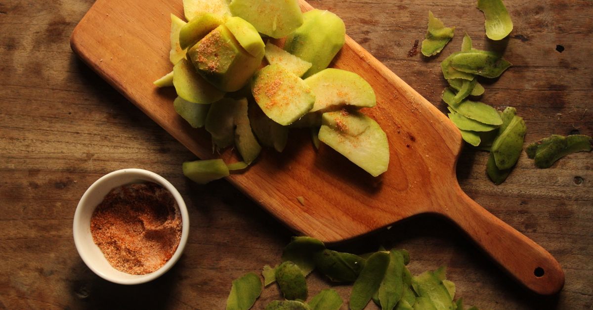 Een houten snijplank met groene fruitschillen en stukjes, met daarnaast een schaaltje kruiden en meer schillen op de houten tafel.