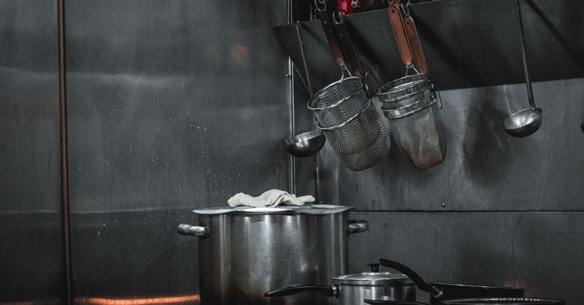Een grote stalen kookpan met een doek op de deksel staat op het fornuis in een professionele keuken, met vergieten en soeplepels eraan opgehangen.