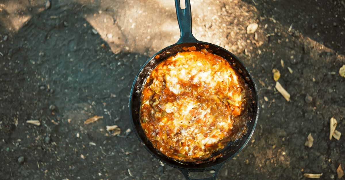 Een gietijzeren koekenpan met een gebakken eiergerecht met tomatensaus, van bovenaf gefotografeerd buiten op de grond.