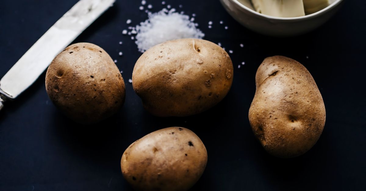 Vier rauwe aardappelen liggen op een donker werkblad naast zout en een schaaltje boter.