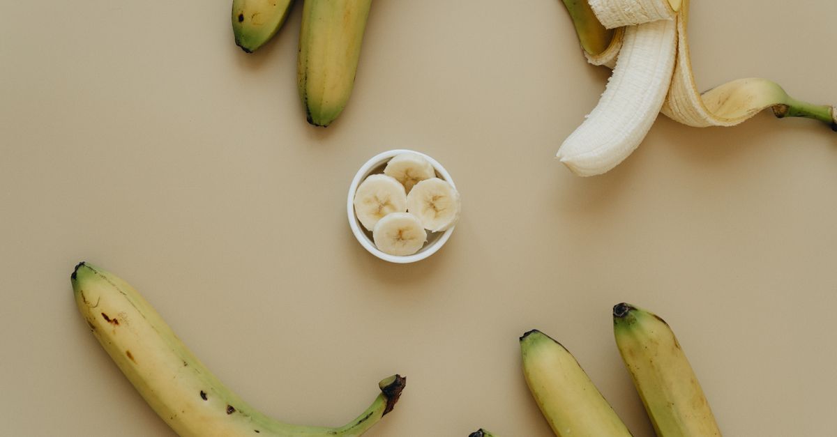 Een klein wit bakje met gesneden bananenschijfjes omringd door hele bananen op een beige achtergrond.