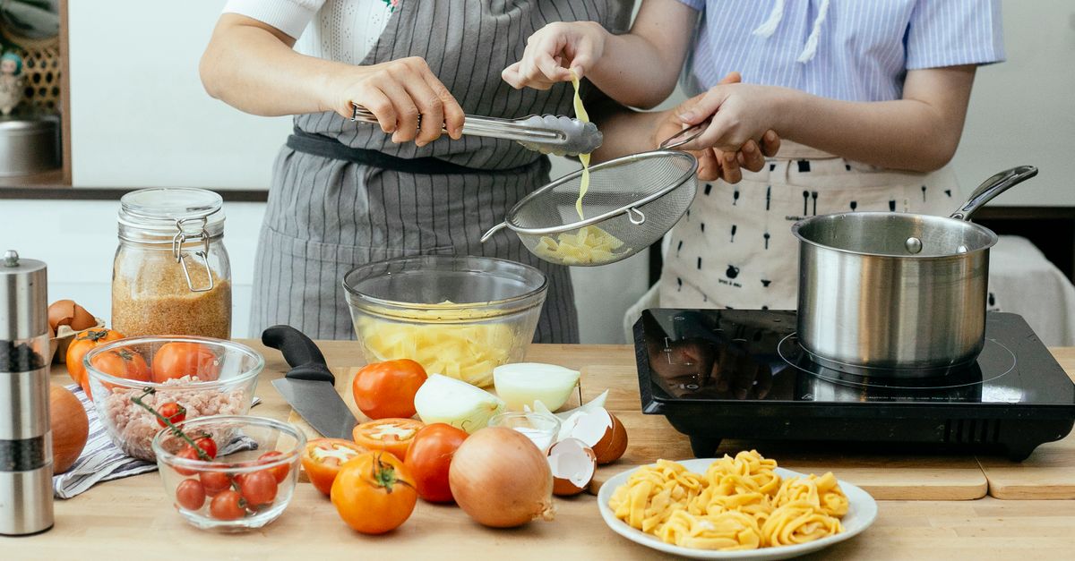Twee personen bereiden pasta in de keuken, waarbij gekookte pasta met een zeef wordt uitgeschept boven een pan op het fornuis, omringd door tomaten en uien.