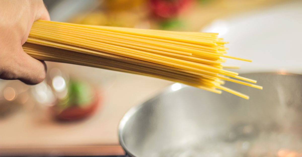 Een hand houdt een bos rauwe spaghetti vast boven een pan met kokend water op het fornuis.