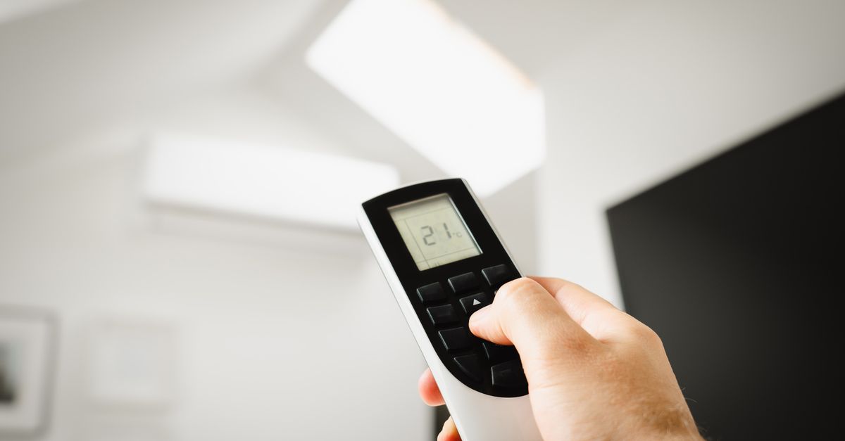 Een hand houdt een afstandsbediening met een temperatuurweergave van 21 graden vast, met een wandunit en een woonkamer op de achtergrond.
