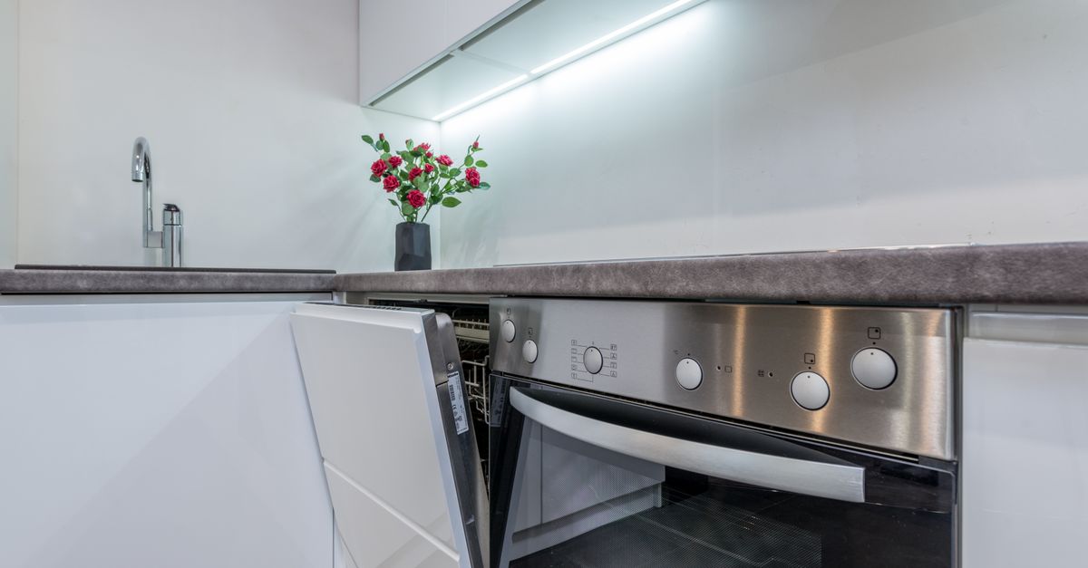 Een geopende inbouwvaatwasser in een moderne witte keuken met een werkblad, kraan en een vaas met rode bloemen.