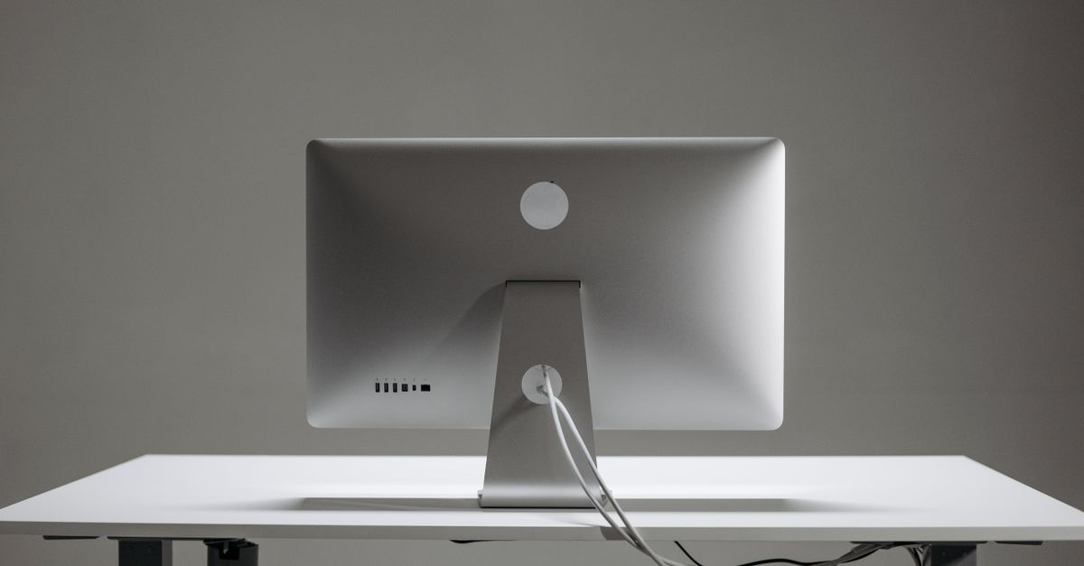 Een minimalistische computermonitor op een wit bureau, gezien van achteren, met slechts één kabel netjes naar beneden lopend.